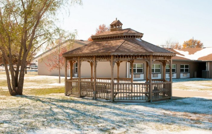 A wooden gazebo in a sunny outdoor setting, surrounded by autumn foliage.