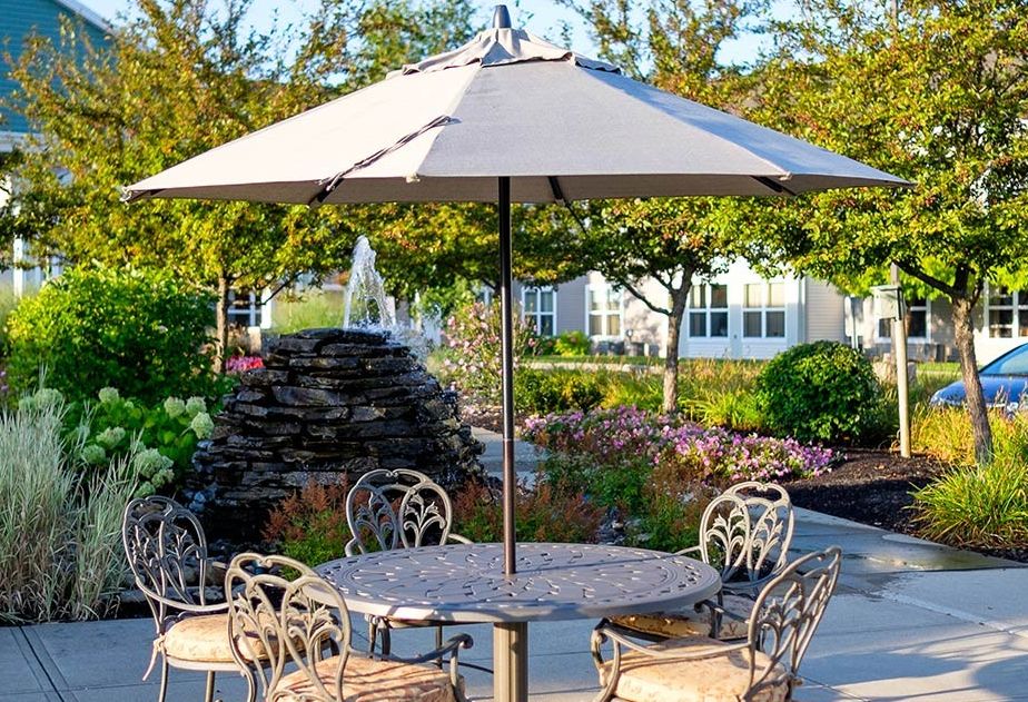 A sunny patio setting with a table, umbrella, and chairs surrounded by lush garden landscaping.