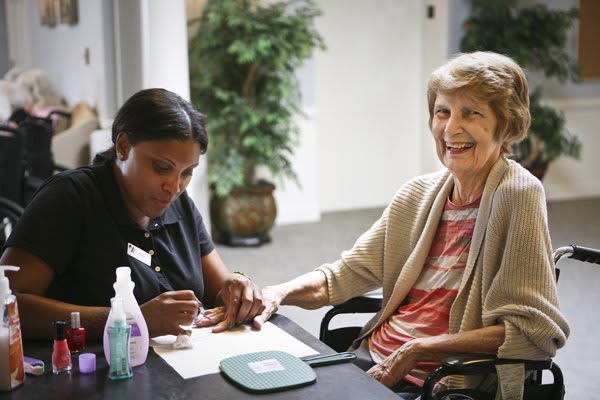 A caregiver gives a manicure to a smiling elderly woman in a comfortable setting.