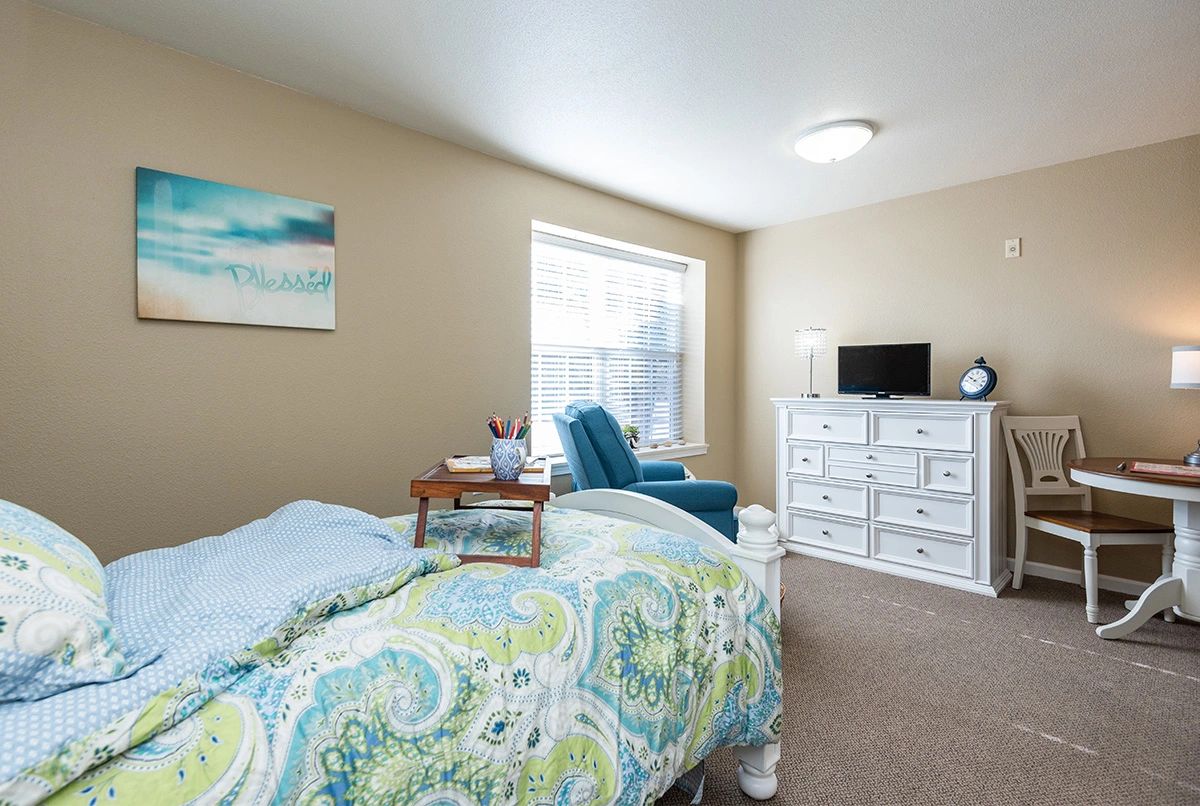 A cozy bedroom with a patterned comforter, armchair, dresser, and a small TV.