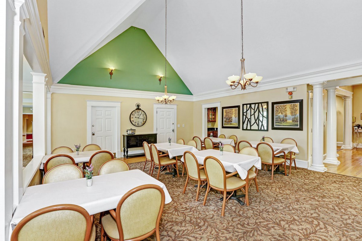 A well-lit dining room with neatly arranged tables and chairs, featuring chandeliers and a green accent wall.