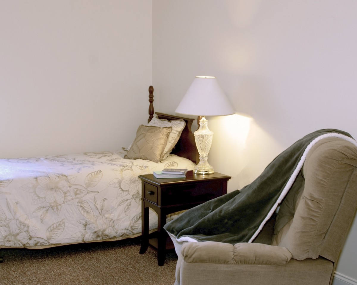 A cozy bedroom with a single bed, nightstand, and lamp, complemented by a comfortable chair and blanket.