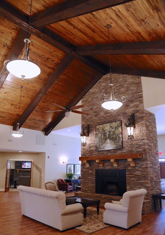A cozy living room with a vaulted ceiling, featuring a brick fireplace and wooden beams, illuminated by chandelier lighting.