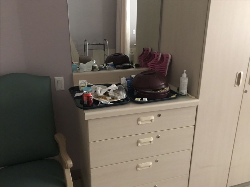 A cluttered dresser with a mirror in a room, featuring various items including a bottle and a chair nearby.