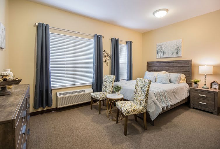 A cozy bedroom with a bed, two chairs, and large windows with curtains.