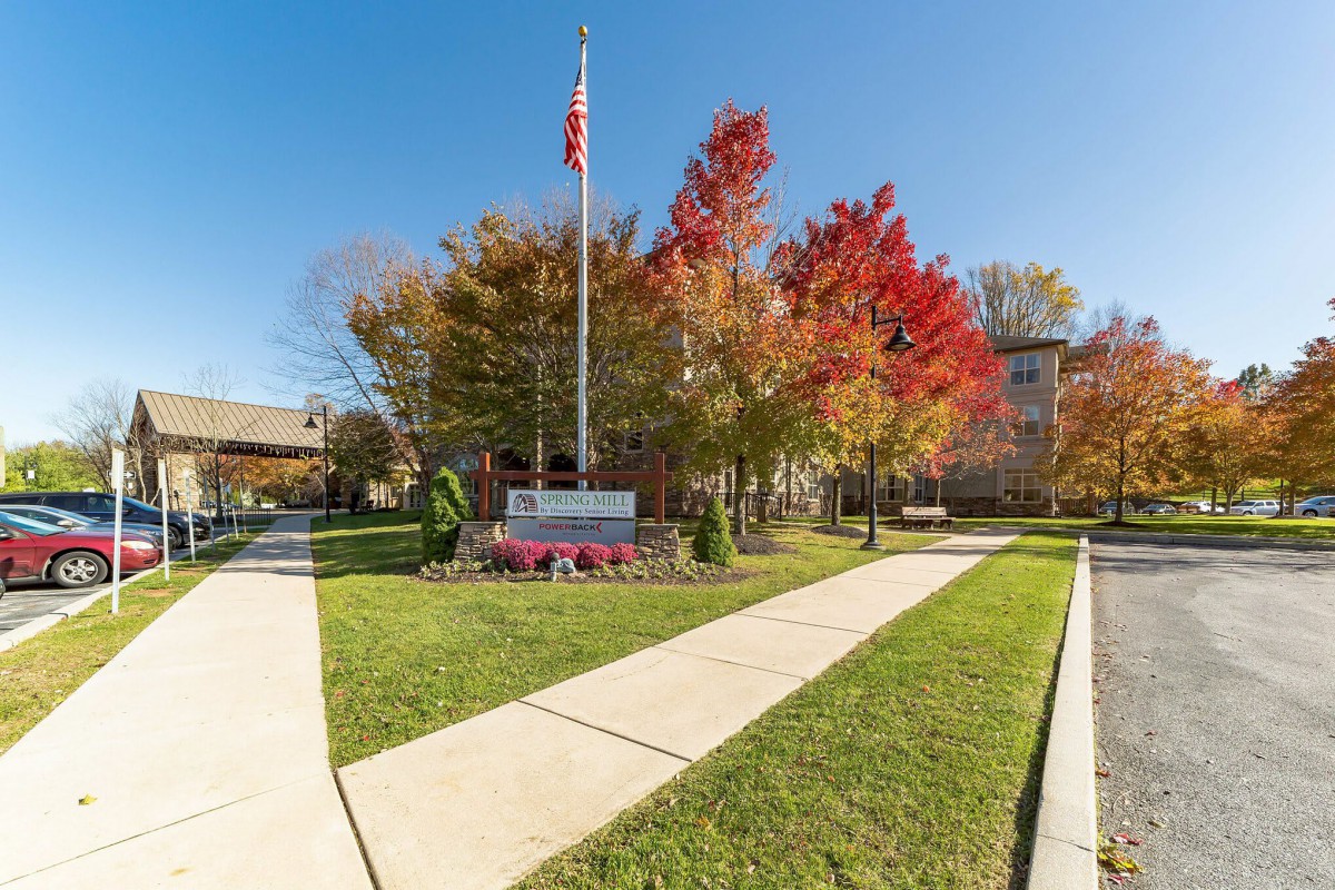 A school entrance with a sign, surrounded by colorful autumn trees and a flagpole.