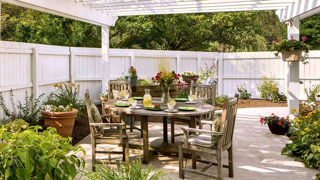 A cozy outdoor patio with a wooden dining set under a pergola, surrounded by lush greenery and potted plants.