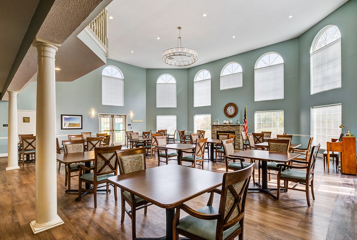 A spacious dining area with multiple tables and chairs, large windows, a chandelier, and wooden flooring.