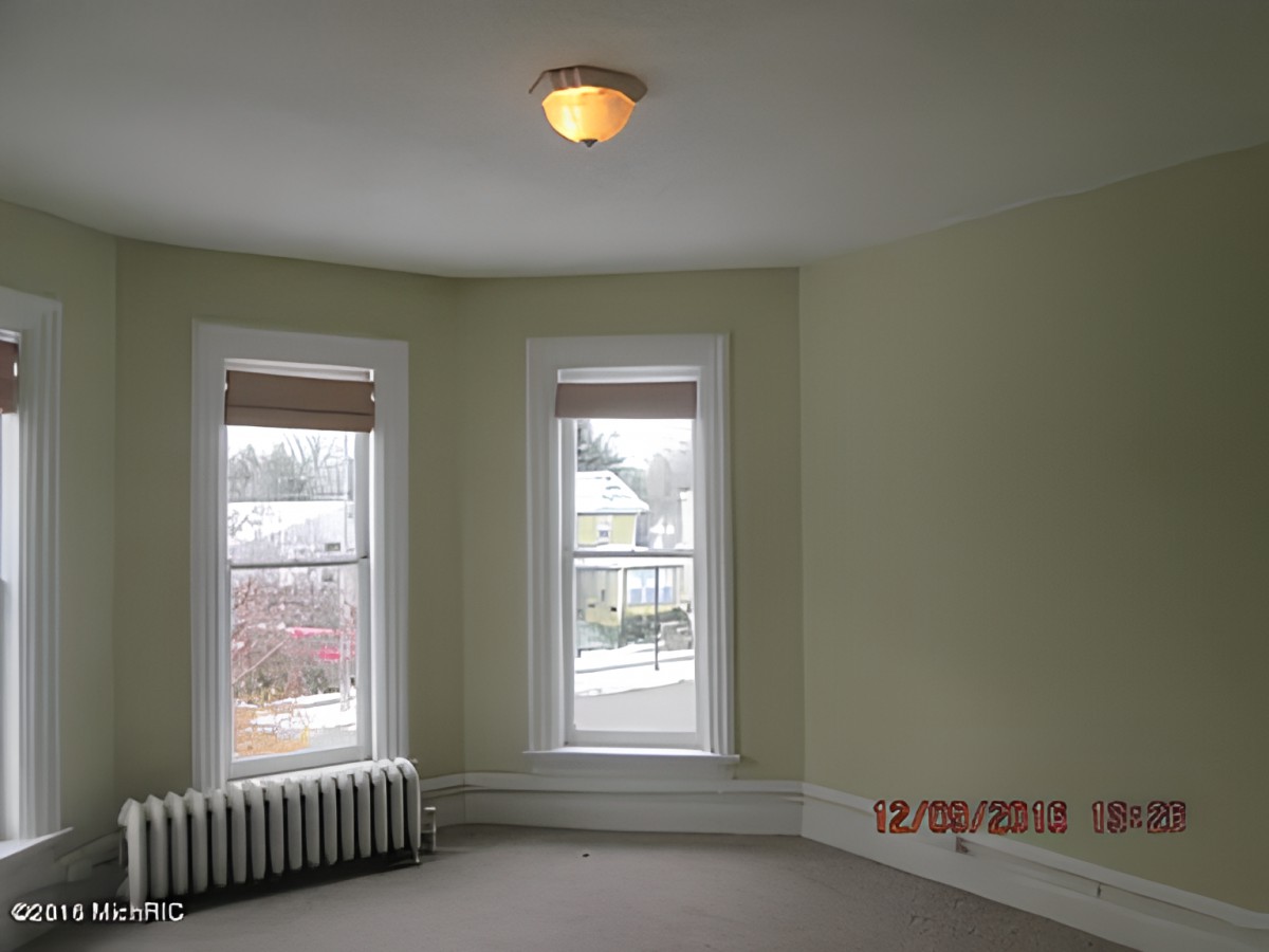 An empty room with carpet flooring, featuring two large windows, a radiator, and a ceiling light.