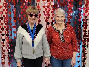Two friends smiling and holding hands in front of a backdrop decorated with red hearts.