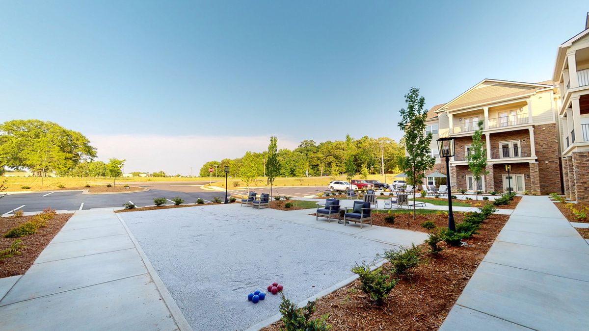 A sunny outdoor patio area with tables and chairs, surrounded by landscaped greenery and a distant view of trees.