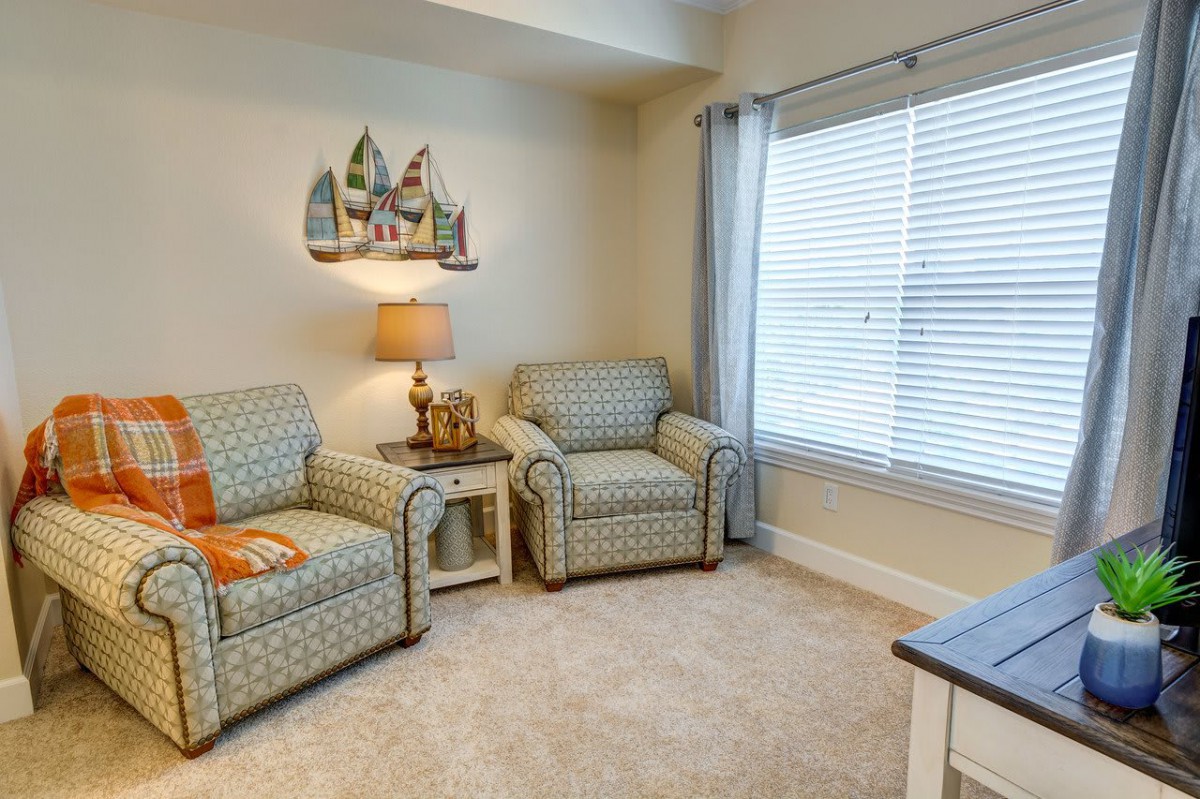 A cozy living room corner with two patterned chairs, a small side table with a lamp, and a window with blinds and curtains.