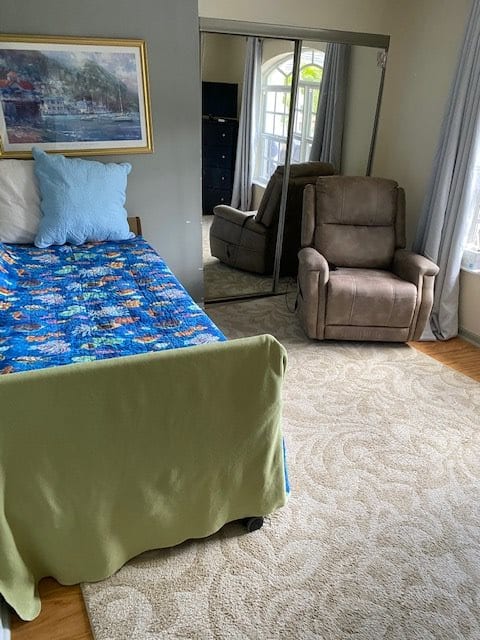 A cozy bedroom with a bed covered in a colorful blanket, a recliner chair, and large windows with curtains.