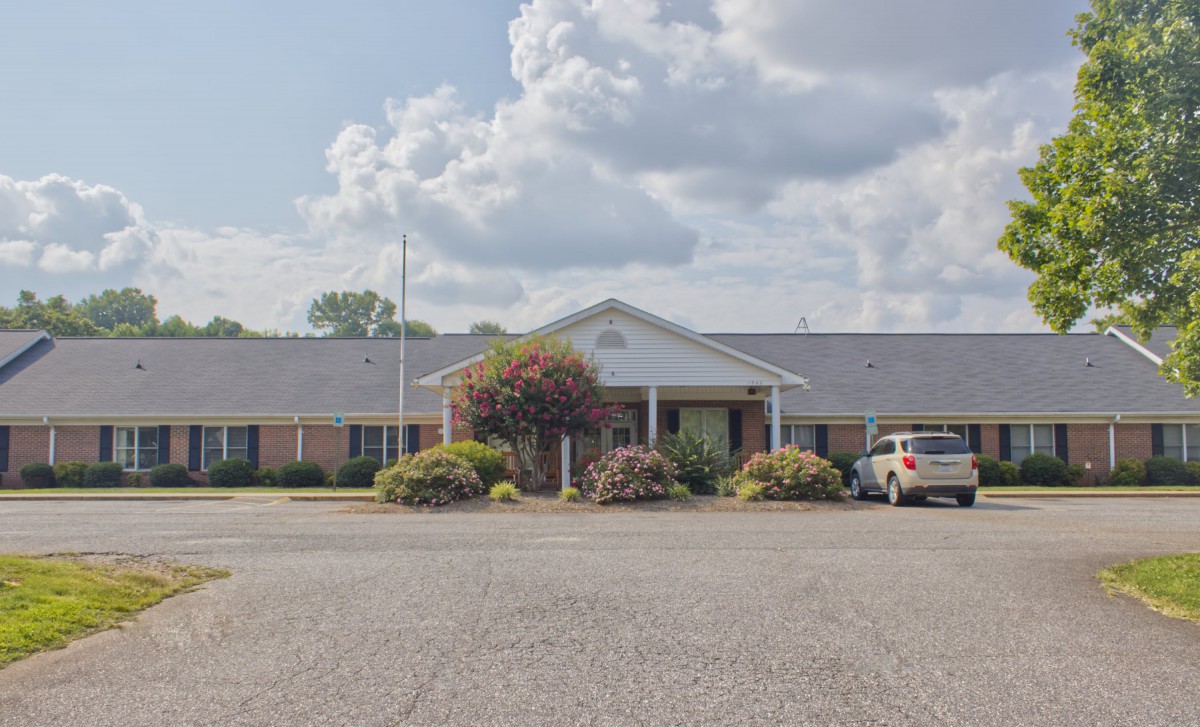A single-story building with a parking lot in front, surrounded by trees under a partly cloudy sky.