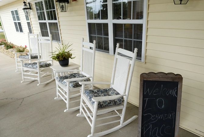 A cozy porch with white rocking chairs, a small table, and a potted plant beside a chalkboard sign.