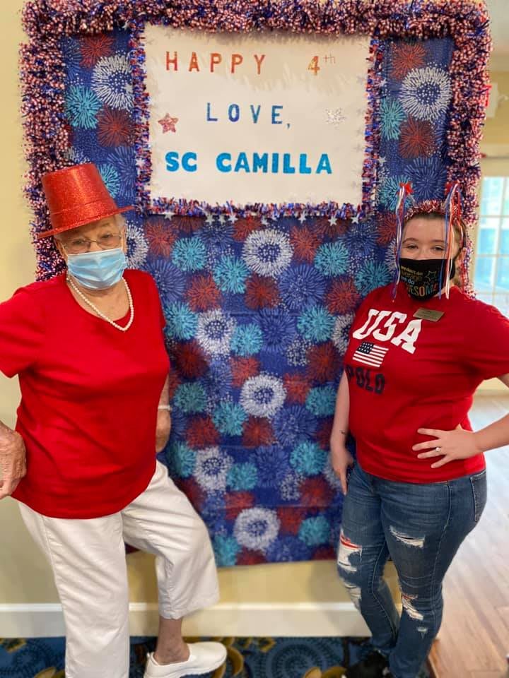Two individuals wearing red shirts and masks pose in front of a decorated backdrop with patriotic colors.