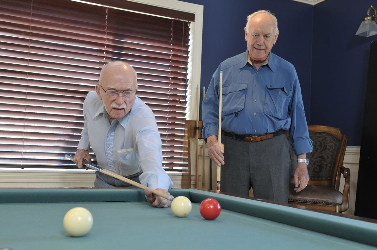 Two older men playing billiards in a room with blue walls and window blinds.