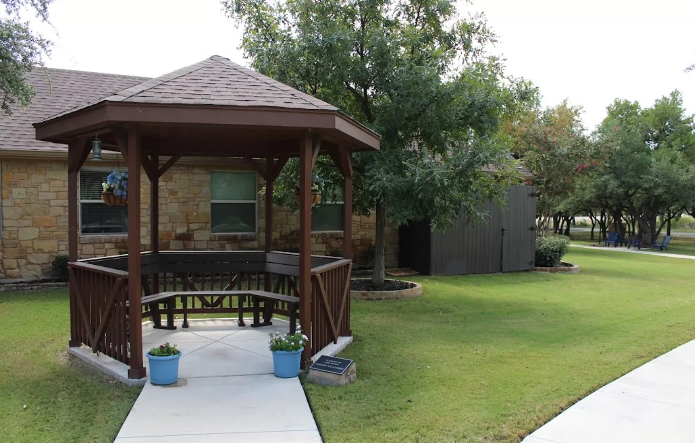 A wooden gazebo in a well-maintained garden setting with lush green grass and trees.