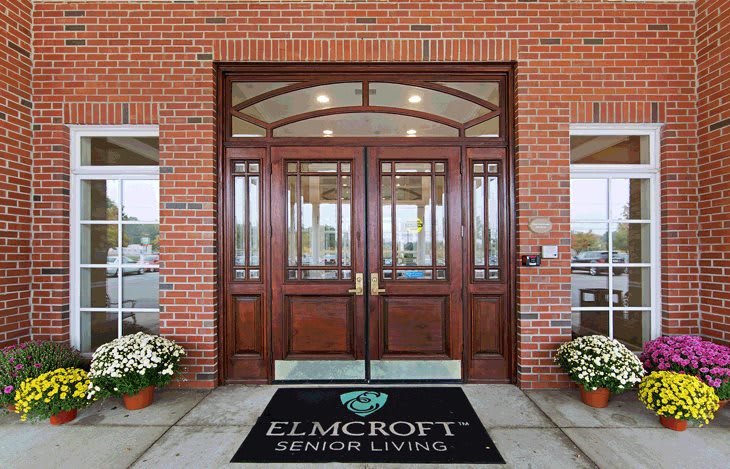Entrance to a senior living facility with wooden doors and a welcome mat.