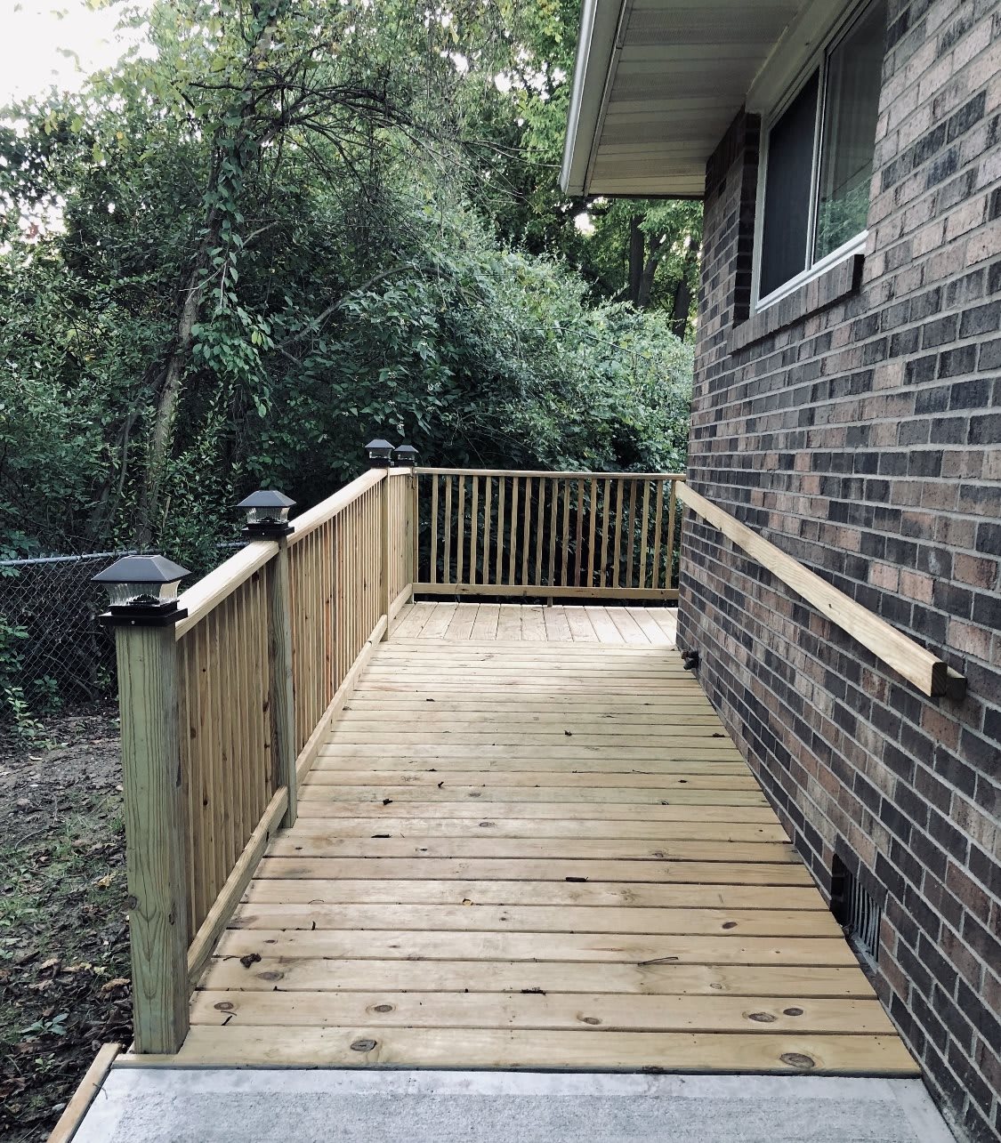 A wooden deck with railings adjacent to a brick house, overlooking a lush backyard.