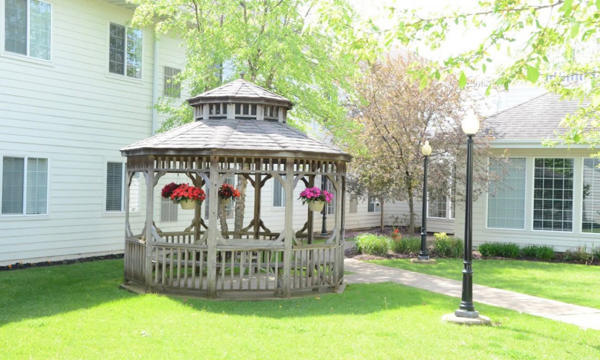 A wooden gazebo adorned with hanging flower baskets, surrounded by lush greenery and lampposts in a garden setting.