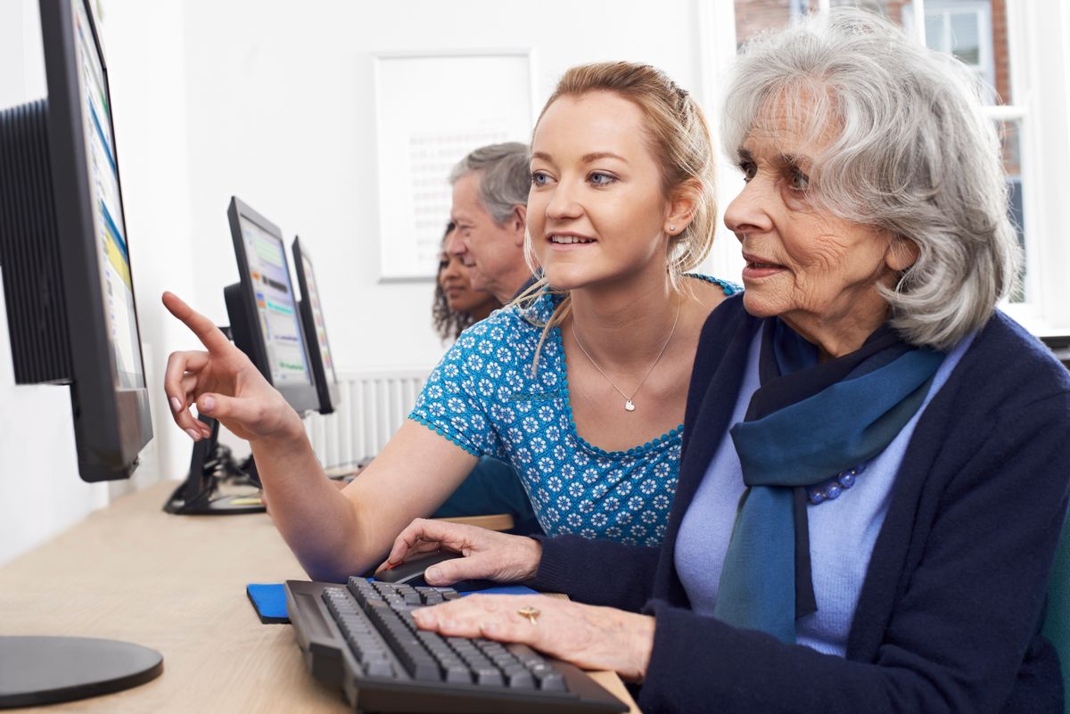 A young woman assists a senior adult with computer use in a classroom setting.
