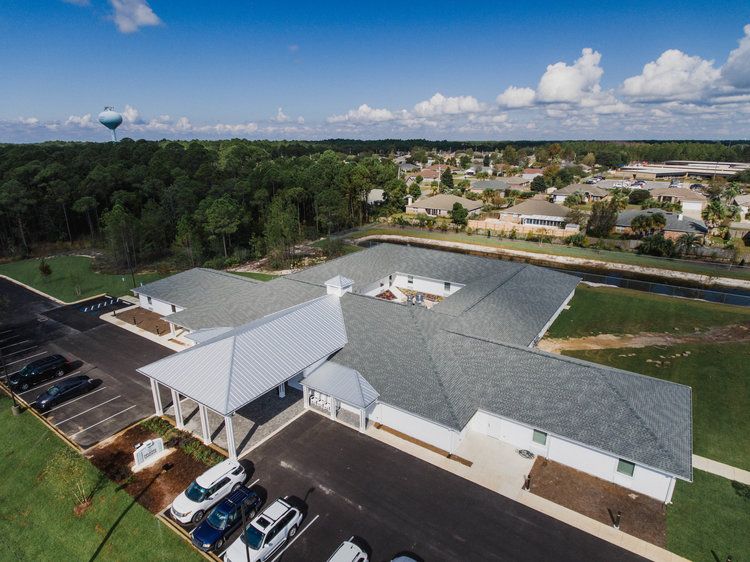 Aerial view of a building complex with a parking lot, surrounded by trees and suburban homes under a blue sky with scattered clouds.