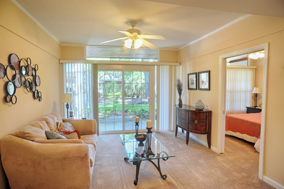 A cozy living room with a sofa, glass coffee table, and ceiling fan, featuring sliding doors that let in natural light.
