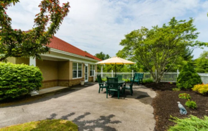 A sunny patio area with a table, chairs, and an umbrella surrounded by greenery.