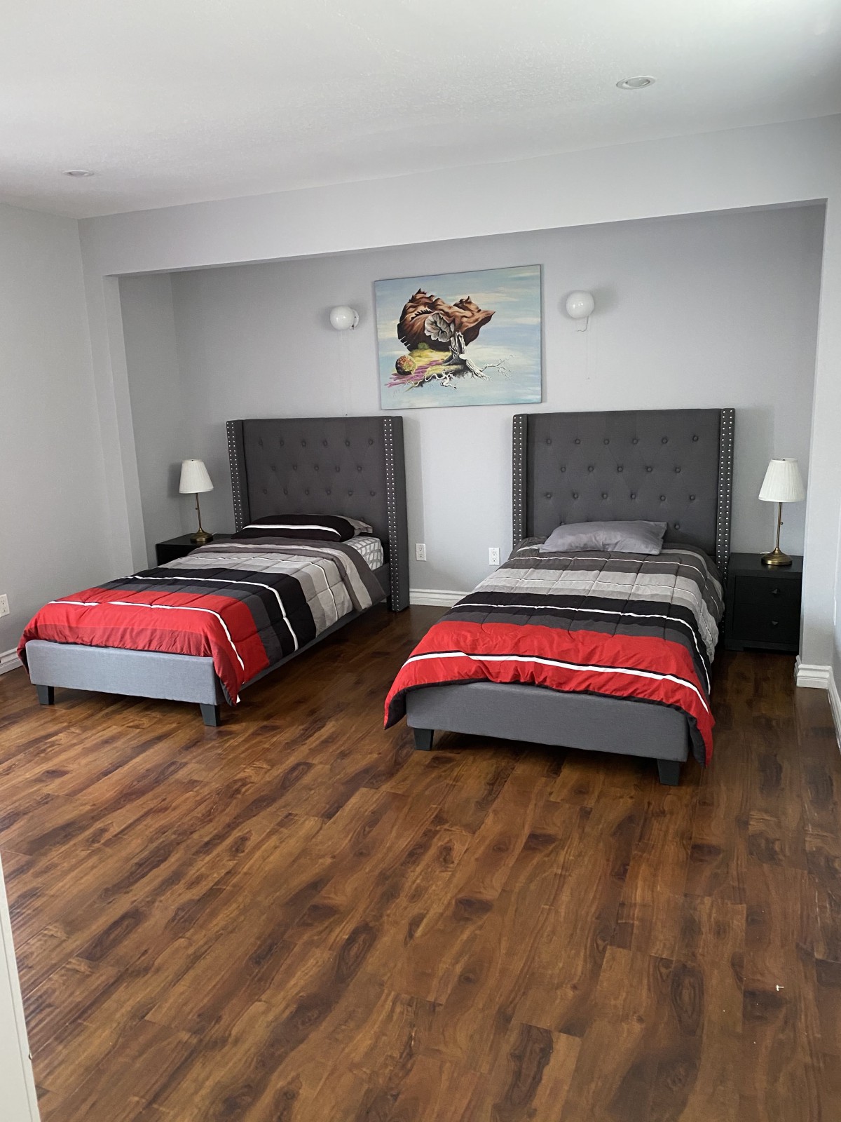 A modern bedroom with two twin beds featuring gray headboards, red and black bedding, lamps on nightstands, and hardwood flooring.