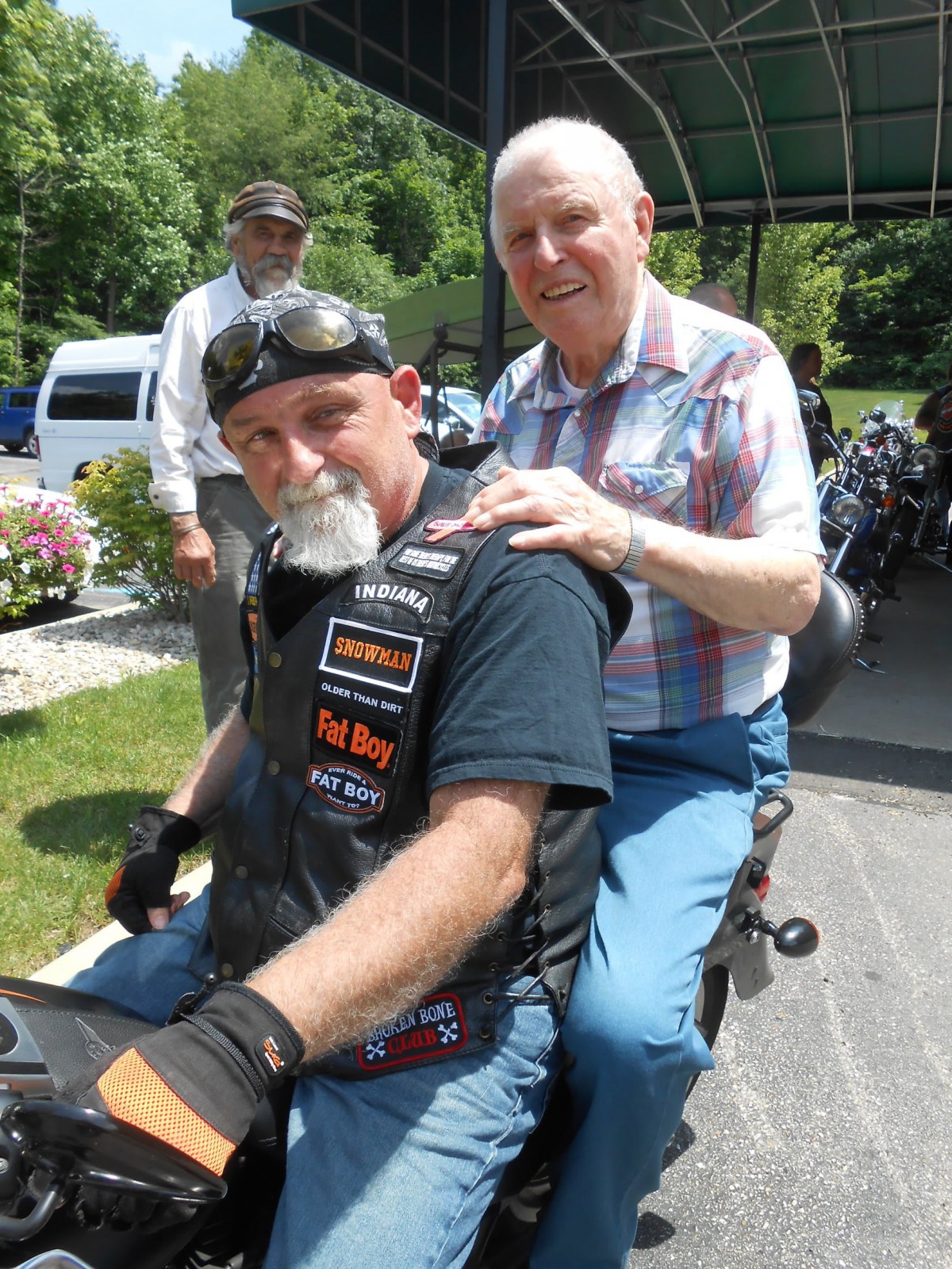 Two men sitting on a motorcycle outdoors, one wearing a leather vest and helmet, the other in casual clothing.