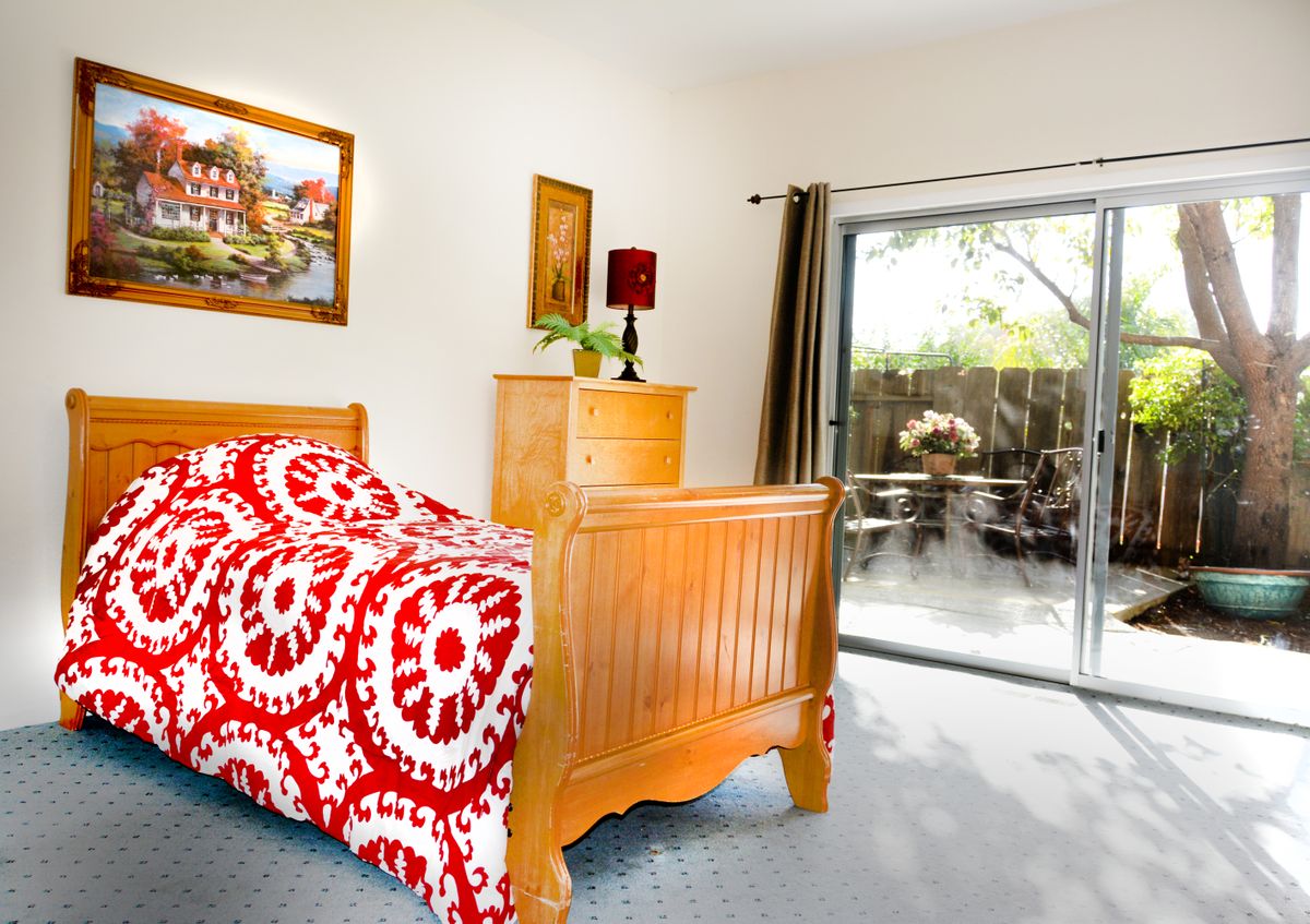 A cozy bedroom with wooden furniture, featuring a bed adorned with a red and white patterned duvet, artwork on the walls, and a sliding door leading outside.