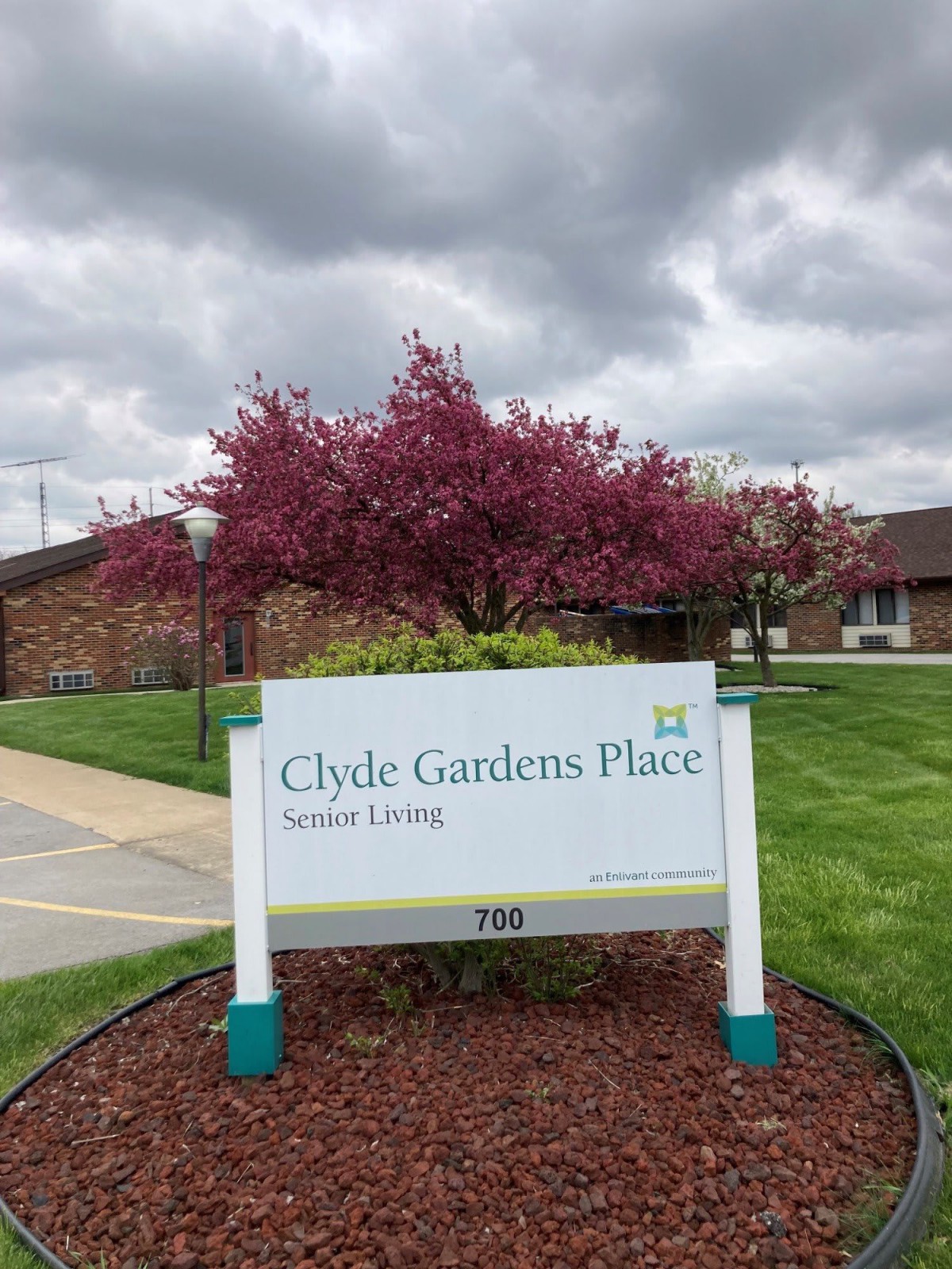 Clyde Gardens Place Senior Living sign in front of a blossoming tree on a cloudy day.