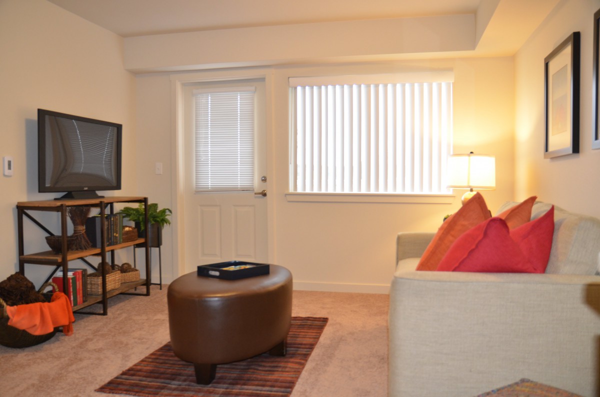 A cozy living room with a sofa, ottoman, and television, illuminated by a floor lamp beside a window with blinds.