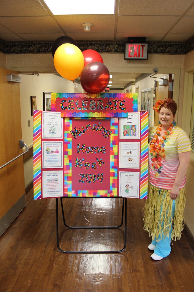 A person dressed in a festive costume stands next to a colorful party poster with balloons attached.
