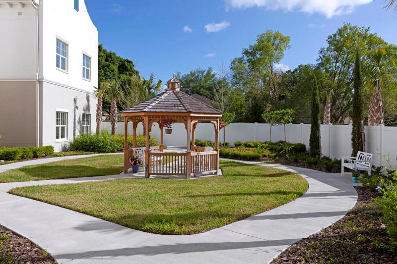 A picturesque gazebo in a garden setting with surrounding pathways, trees, and a nearby building.