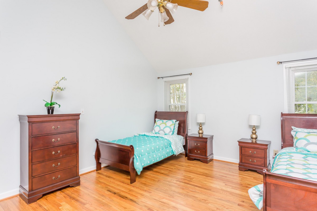 A simple bedroom with wooden furniture, including a bed, nightstands, and a dresser, featuring hardwood floors and a ceiling fan.