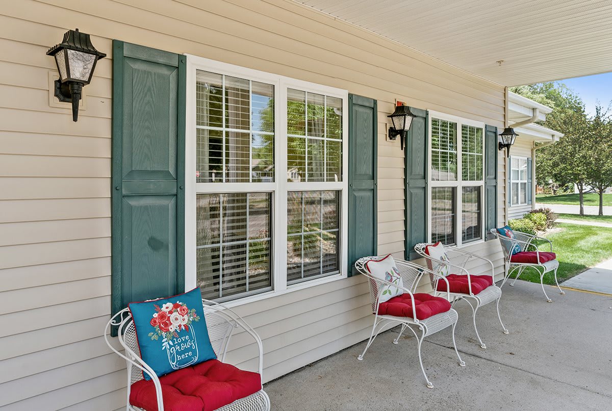 A cozy porch with white chairs and red cushions beside a window with green shutters.