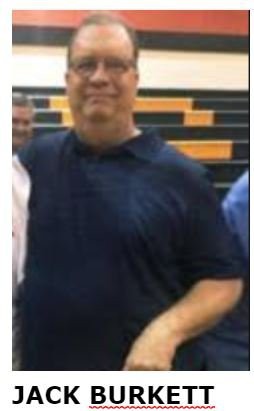 A person in a navy blue polo shirt standing indoors with a gymnasium background.