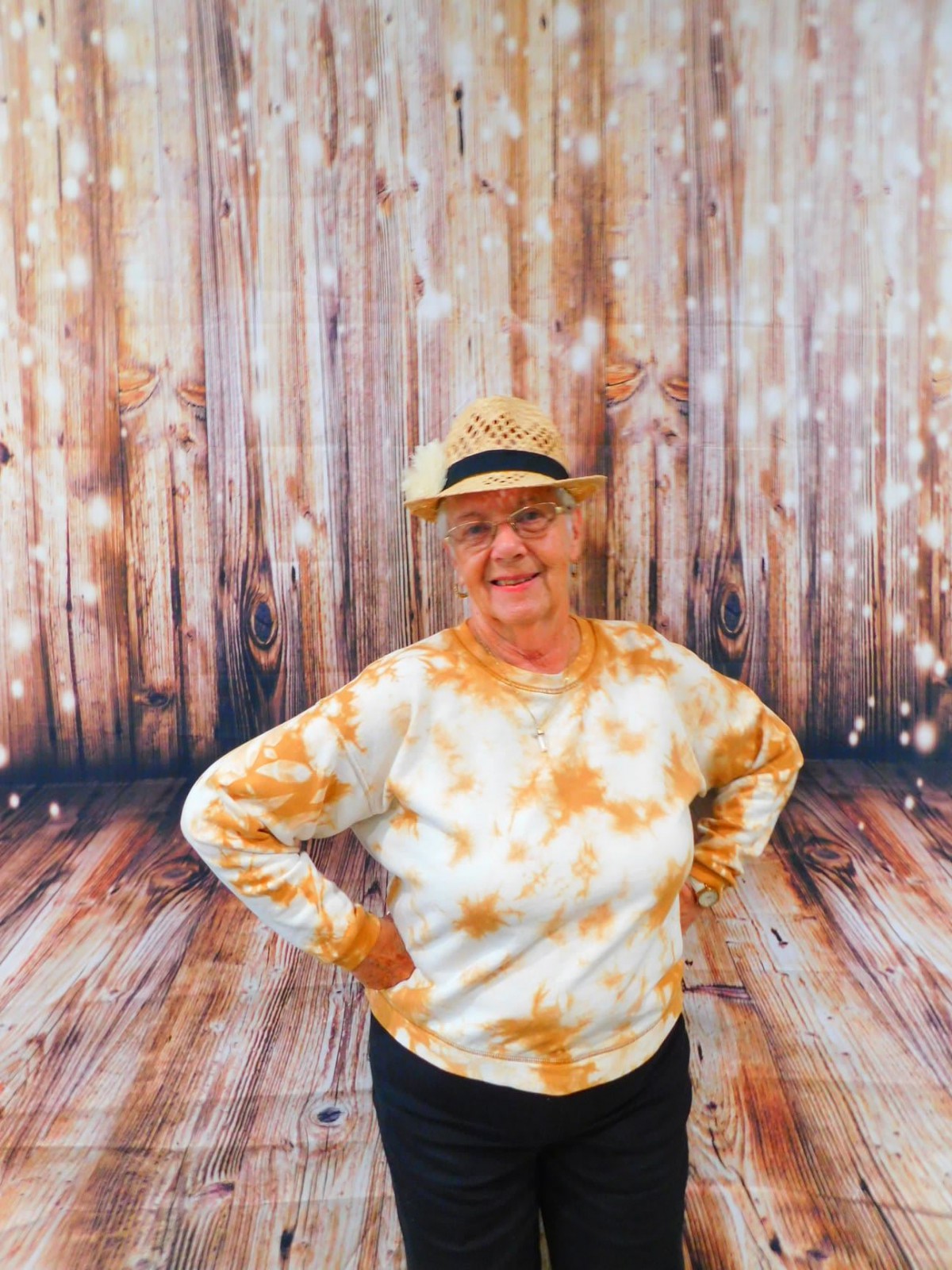 A person smiling while wearing a hat and casual tie-dye clothing, standing in front of a wooden backdrop.