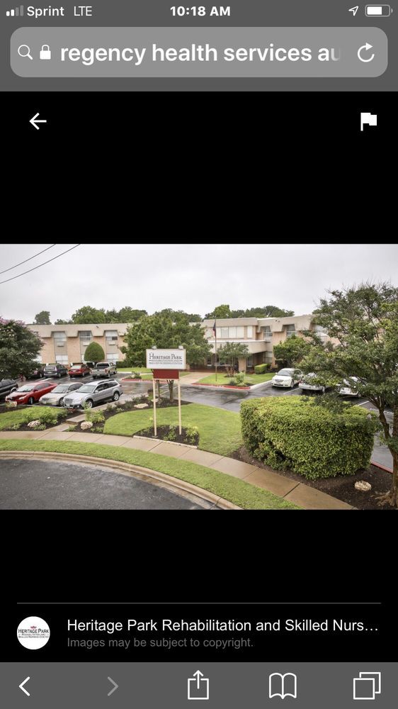 An apartment complex with a parking lot and several parked cars, surrounded by greenery under an overcast sky.