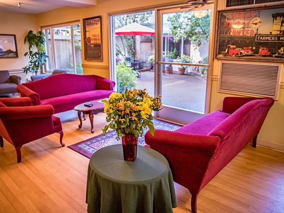 A cozy living room with red sofas and a vase of flowers, illuminated by natural light through glass doors.