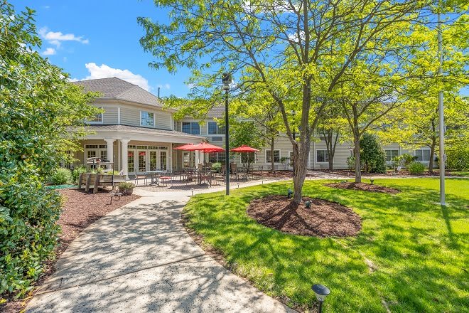 A sunny day with a view of a well-maintained building surrounded by trees and landscaped gardens, featuring a pathway and patio area with red umbrellas.