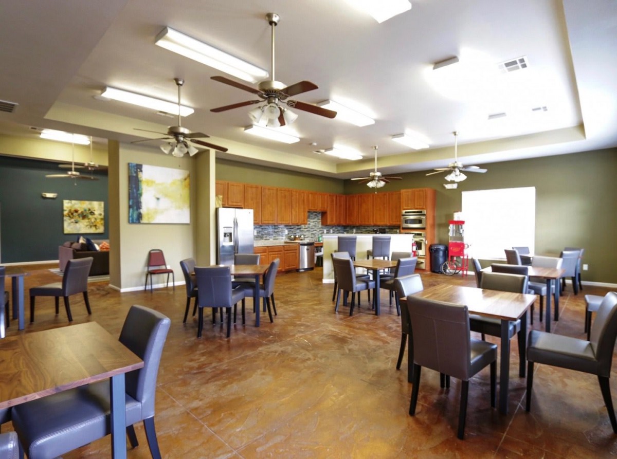 A spacious dining area with multiple tables and chairs, featuring a kitchen area and ceiling fans.
