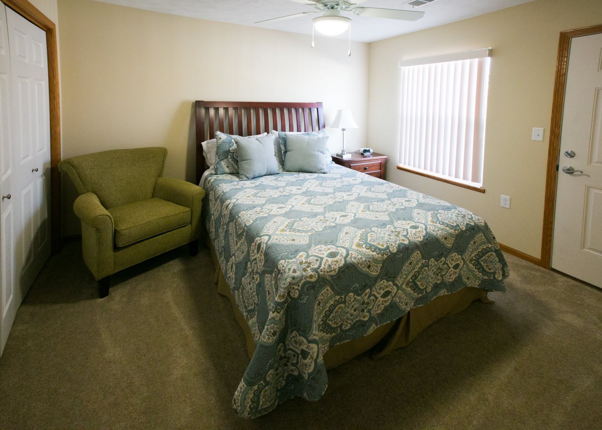 A cozy bedroom featuring a bed with floral bedding, an armchair, and a bedside table with a lamp.