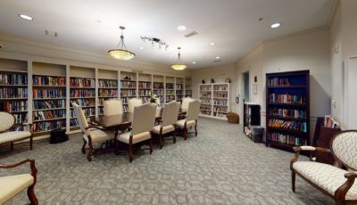 A cozy library room with a large table surrounded by chairs, filled with shelves of books and soft lighting.