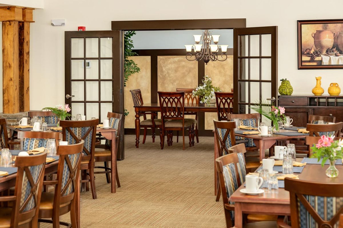 A cozy dining room with neatly arranged tables and chairs, featuring decorative plants and a chandelier.