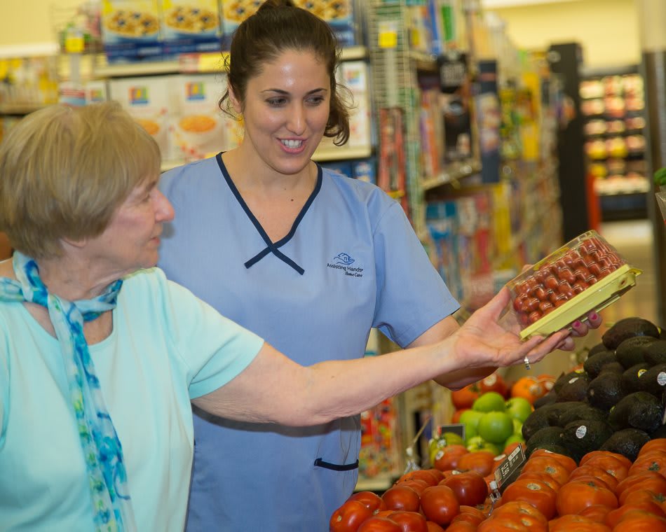 An elderly woman receives assistance from a caregiver while shopping for produce in a grocery store.