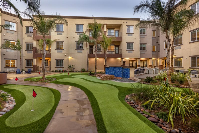 A residential apartment complex with a landscaped garden and pathway, surrounded by palm trees.