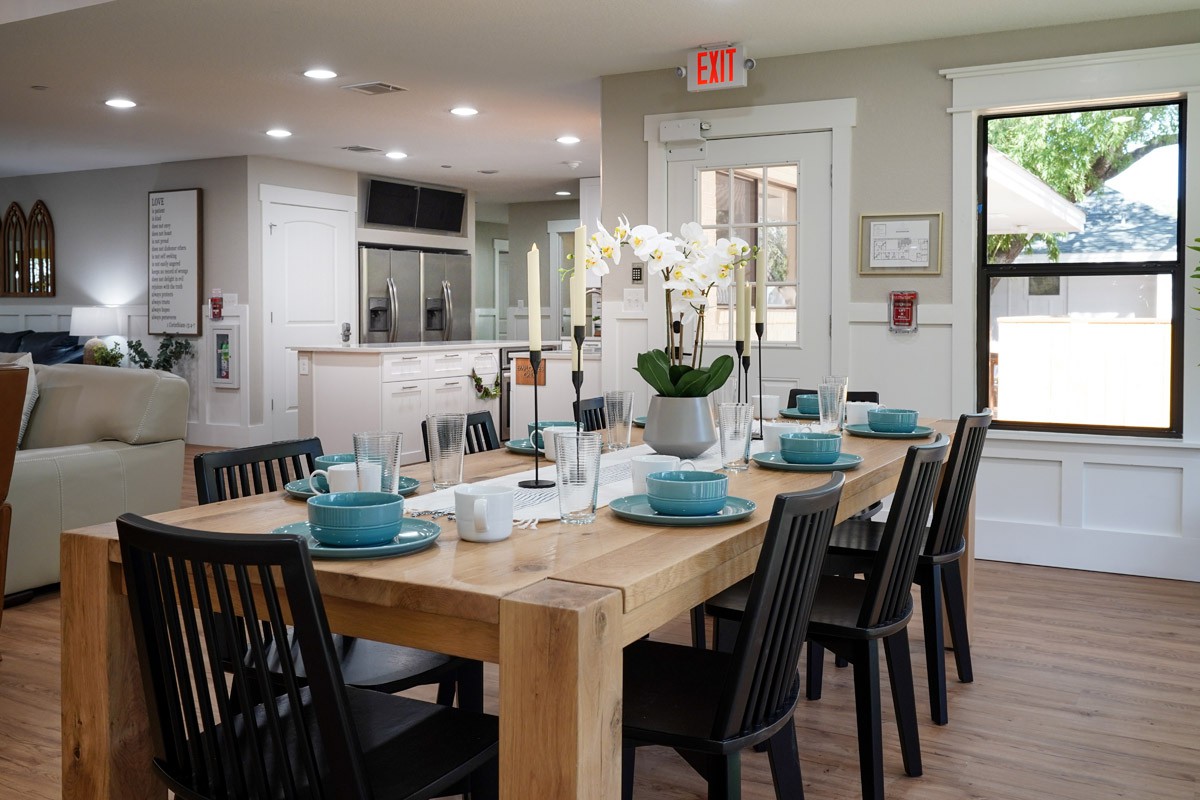 A modern dining room with a wooden table elegantly set with teal dishes and an orchid centerpiece, adjacent to an open kitchen.
